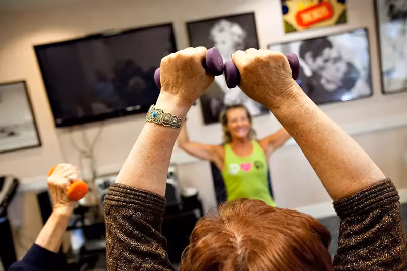 Two elderly individuals lifting small dumbbells during a fitness class led by an instructor in a room with framed pictures and a television on the wall.
