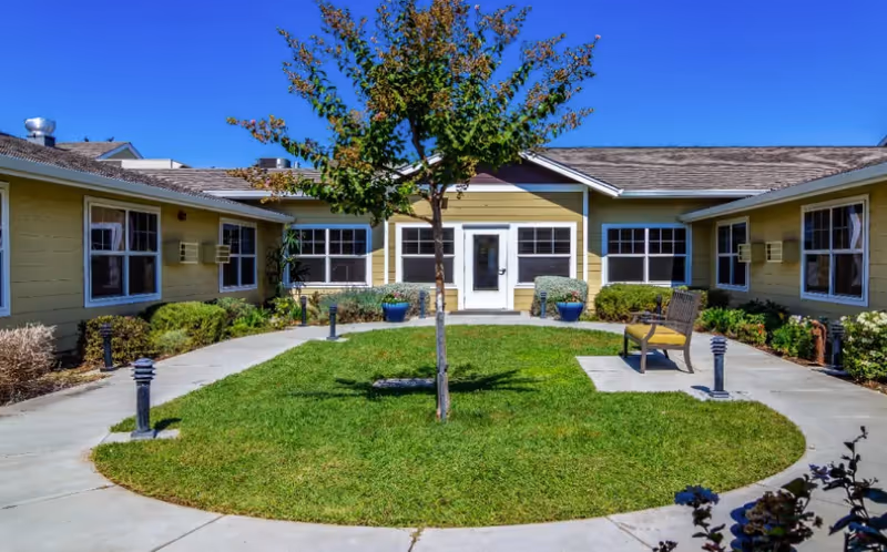 A courtyard area at Amber Grove Place Memory Care featuring a circular grassy lawn with a small tree in the center, surrounded by a concrete walkway. The courtyard is enclosed by a single-story yellow building with multiple windows and a white door. There are benches and potted plants along the walkway under a clear blue sky.