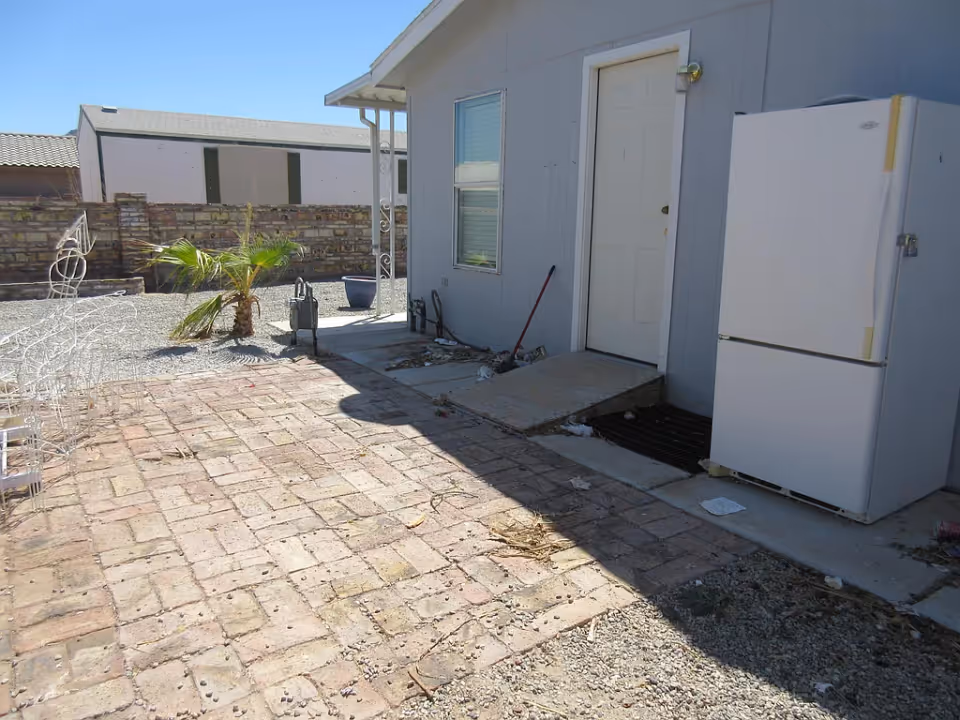 Outdoor area with a brick-paved patio and gravel ground. There is a small palm plant near a brick wall, a white refrigerator placed outside next to a light gray building with a white door and window. Some scattered debris and a broom are visible near the door.
