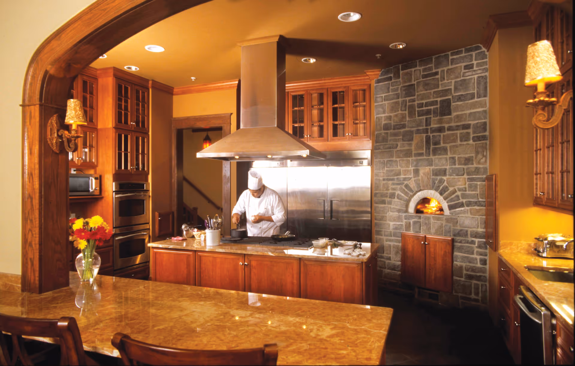 A chef in a white uniform and hat is cooking in a warm-toned kitchen with wooden cabinets, a large stainless steel refrigerator, a stone wall with a small wood-fired oven, and a marble countertop island with utensils and a vase of flowers.