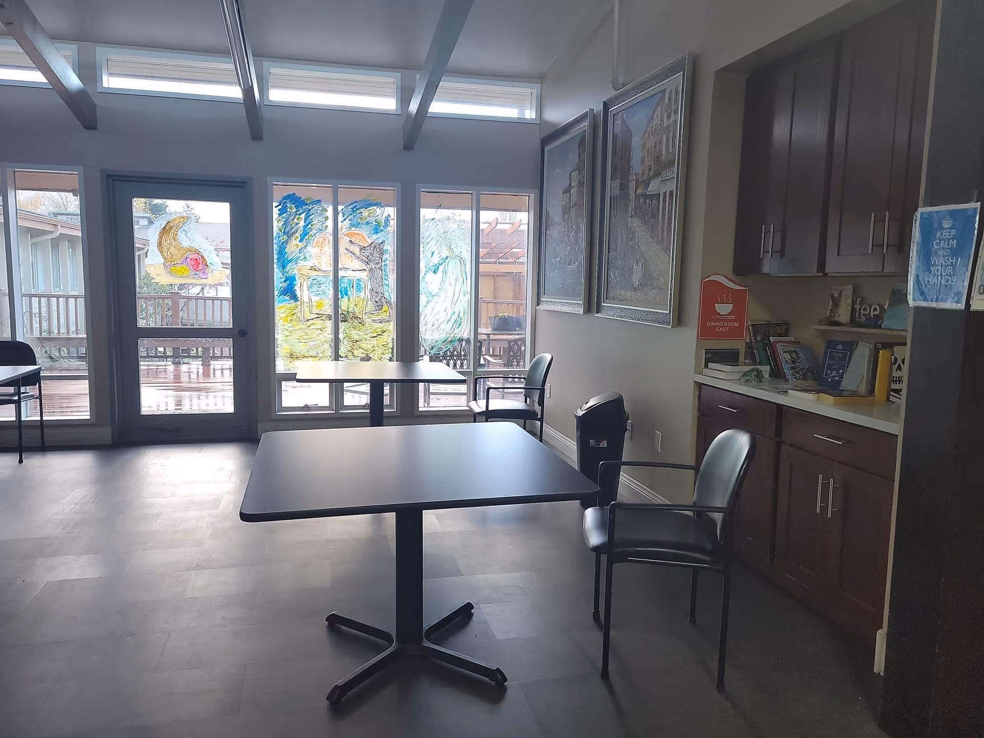 A dining area with two black tables and black chairs. The room has large windows and a glass door with colorful drawings on them. There are two framed pictures on the wall and a cabinet with books and decorations. A sign on the wall reads 'Dining Room East' and another sign reminds to keep calm and wash hands.