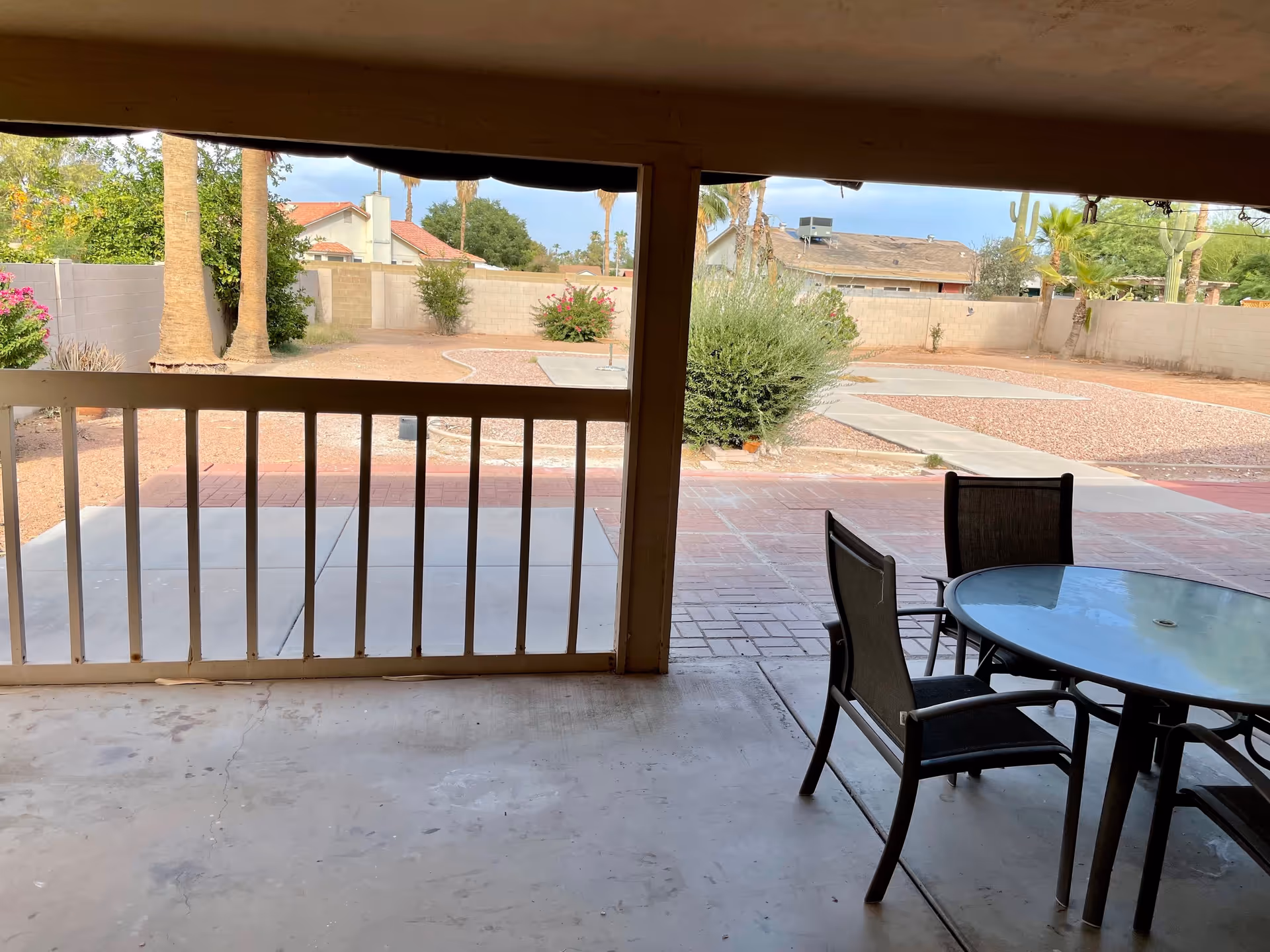 View from a covered patio area looking out onto a backyard with a concrete floor, a round glass table with three chairs, a white railing, and a landscaped yard with bushes, palm trees, and a brick wall fence.