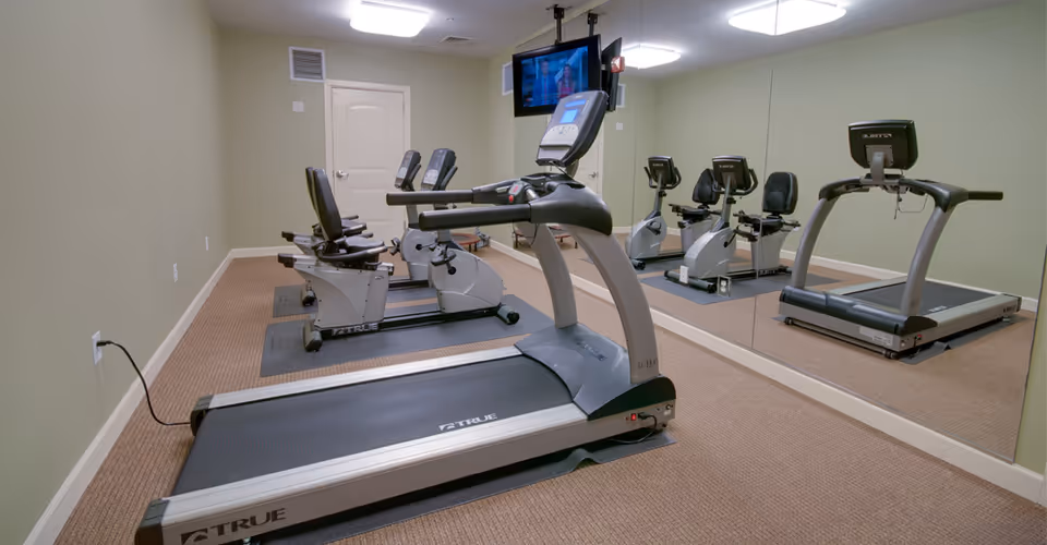 A small fitness room with exercise equipment including a treadmill and two recumbent stationary bikes. The room has beige carpet, light green walls, a large wall mirror, and a wall-mounted TV.