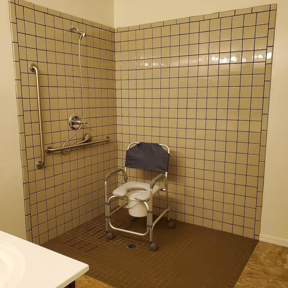 Accessible tiled shower area with grab bars and a wheeled commode/shower chair.