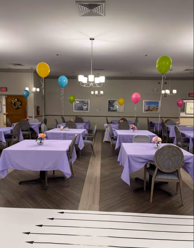 Dining room with multiple tables draped in lavender tablecloths, floral centerpieces, and colorful balloons tied to chairs.