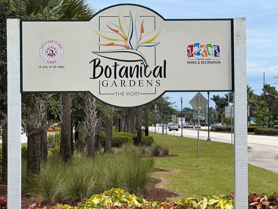 White sign reading "Botanical Gardens — The Port" mounted on posts in a grassy roadside planting with palm trees and a street in the background.