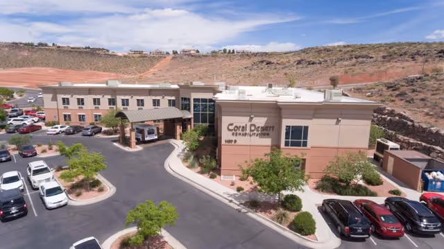 Aerial view of Coral Desert Rehabilitation And Care facility showing a large building with a covered entrance, surrounded by parking lots with several cars parked. The building is situated in a desert-like landscape with hills and sparse vegetation in the background under a partly cloudy sky.