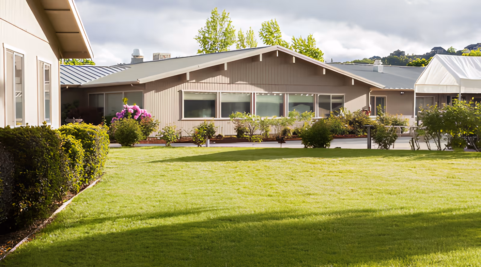 A well-maintained outdoor garden area at Royale Gardens Health & Rehabilitation Center with green grass, bushes, and flowering plants in front of a single-story building with large windows and a gray roof under a cloudy sky.