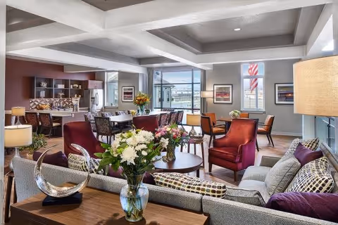 A spacious and well-lit common area in an assisted living facility featuring a modern kitchen with a breakfast bar and stools, a dining table with chairs, and a comfortable seating area with sofas and armchairs. The room is decorated with fresh flowers, artwork on the walls, and large windows letting in natural light.