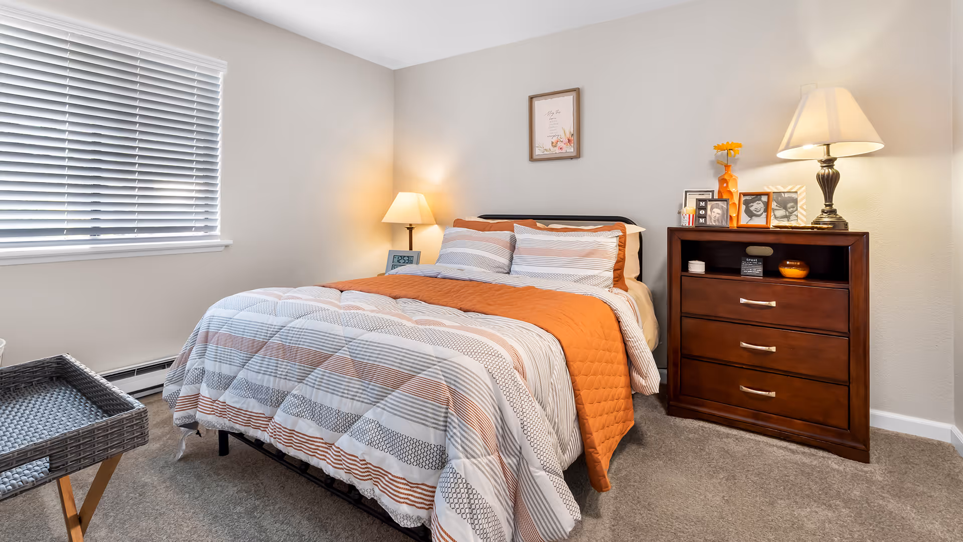 A neatly made bedroom with orange-accented bedding, a bedside lamp, and a wooden dresser topped with framed photos.
