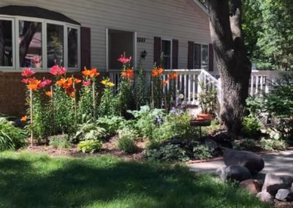 A garden area in front of a residential building with colorful flowers including orange, pink, and yellow blooms, green plants, a large tree, and a white railing along a porch or deck.