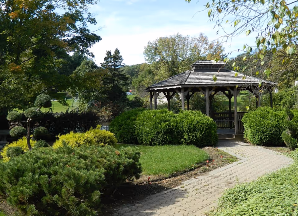 A peaceful outdoor garden area with a wooden gazebo surrounded by green bushes, trees, and a paved walkway leading to the gazebo under a clear blue sky.