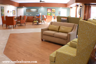 A spacious and well-lit living room area in a senior living facility with comfortable seating including a beige loveseat with two green cushions, green patterned armchairs, and several tables and chairs in the background. The room has a mix of wood and tile flooring, wall art, and large windows with curtains allowing natural light to enter.