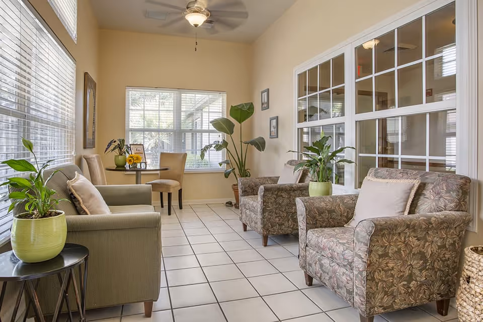Sunlit seating area with upholstered armchairs, potted plants, and a small table in a senior living lounge.