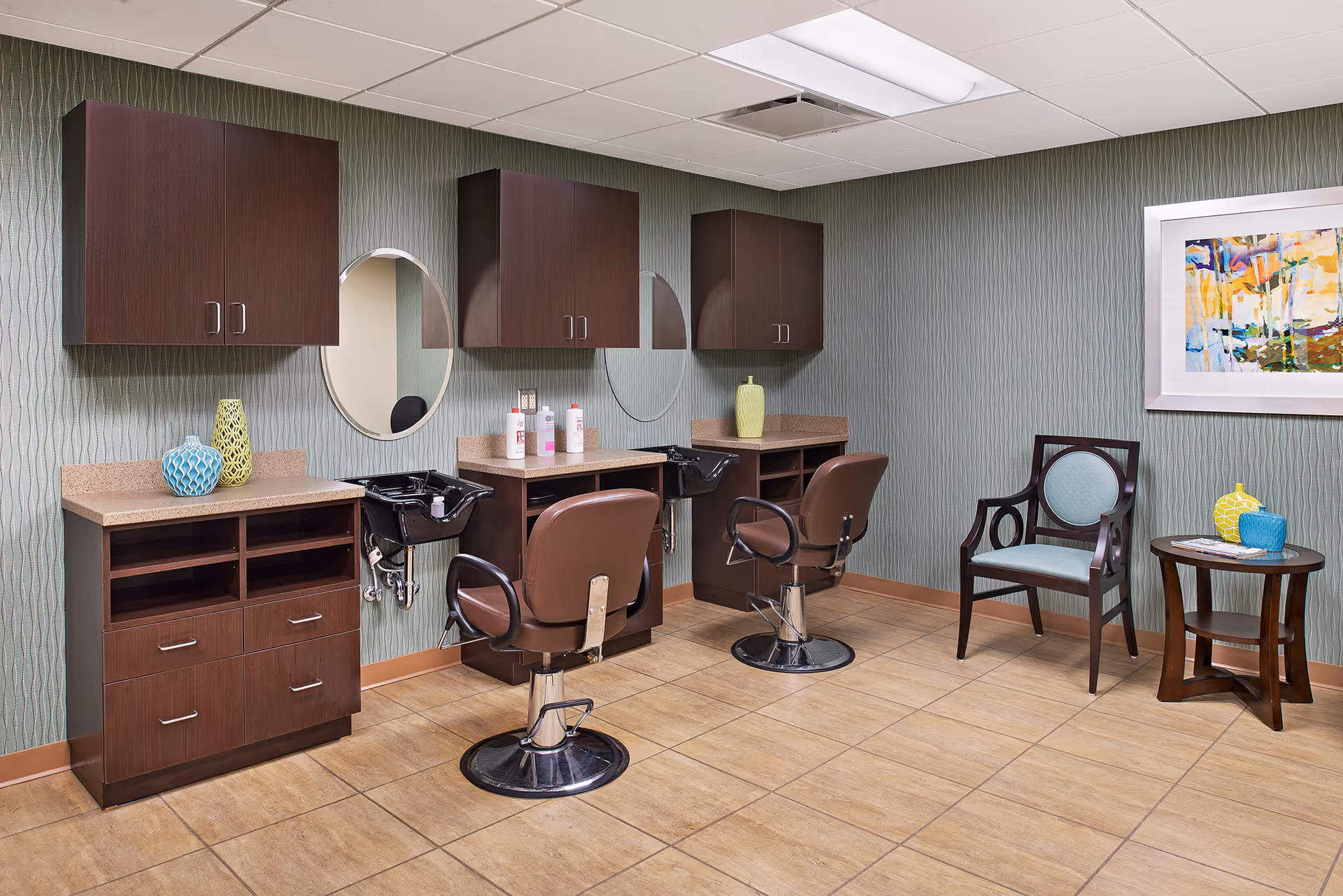 Interior view of a salon area in a senior living facility with two brown salon chairs in front of two wash basins and mirrors. There are dark wood cabinets mounted on the wall and a matching cabinet below the counter. To the right, there is a wooden chair with light blue upholstery and a small round wooden table with decorative vases and magazines. A colorful abstract painting is hung on the wall.