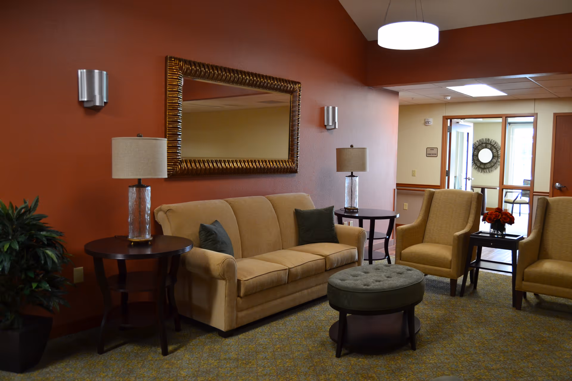 Comfortable lobby-style seating area with a beige sofa, armchairs, side tables, lamps, ottoman, a large mirror and a potted plant against an orange wall.