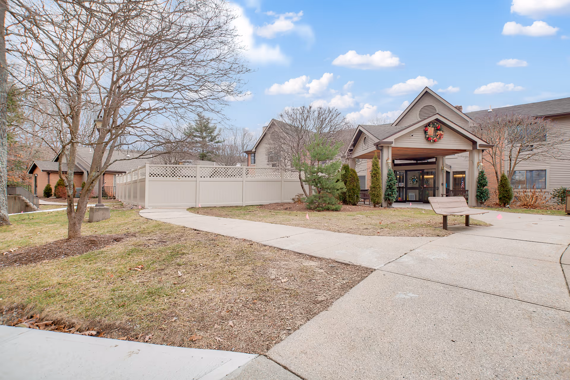 Exterior view of a senior living facility entrance with a covered porch decorated with a holiday wreath. There is a paved walkway leading to the entrance, a bench on the right side, leafless trees, and a fenced area on the left. The sky is partly cloudy.