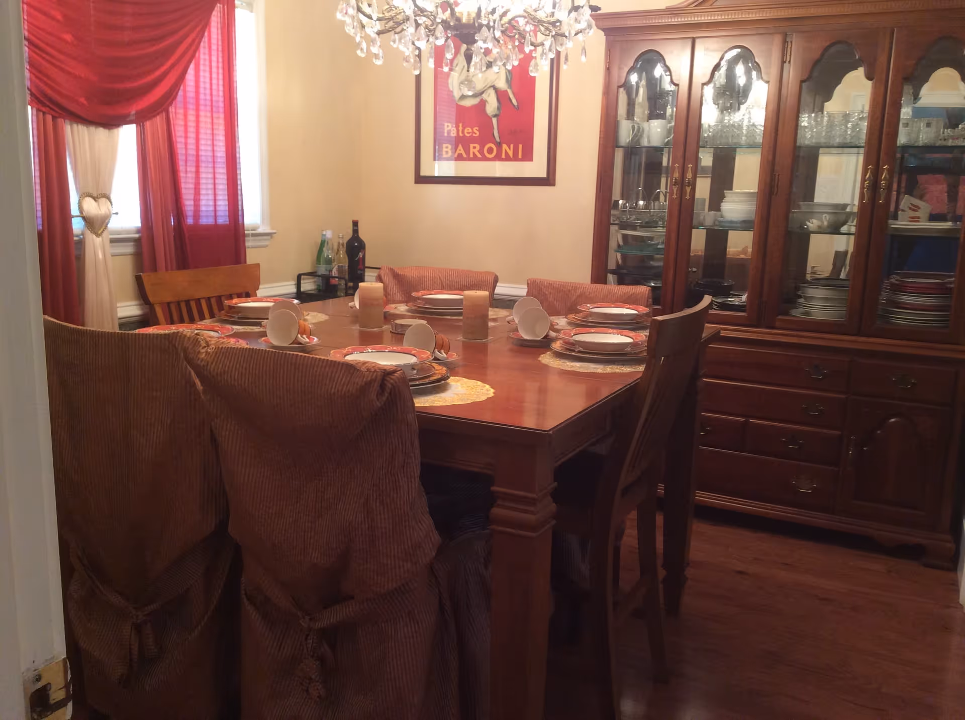 A dining room with a wooden dining table set for six people. The table has plates, bowls, and candles arranged on placemats. There are six chairs around the table, two of which have brown slipcovers. A wooden china cabinet filled with dishes and glassware is against the wall. Red and cream curtains cover the windows, and a chandelier hangs above the table. A framed poster is on the wall behind the table.
