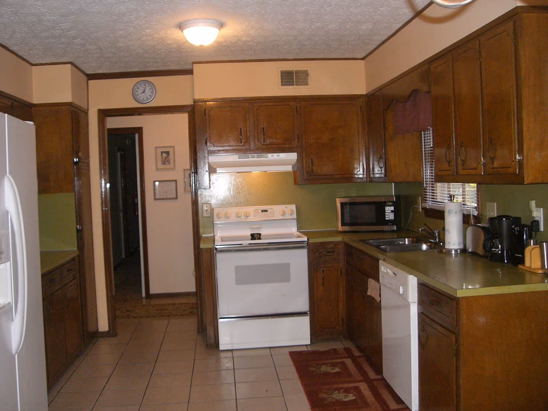 Interior view of a kitchen with wooden cabinets, a white stove with a range hood, a microwave, a dishwasher, a refrigerator, a coffee maker, and a sink. The floor is tiled, and there is a red patterned rug in front of the stove and sink. A clock is mounted above the doorway leading to a hallway.