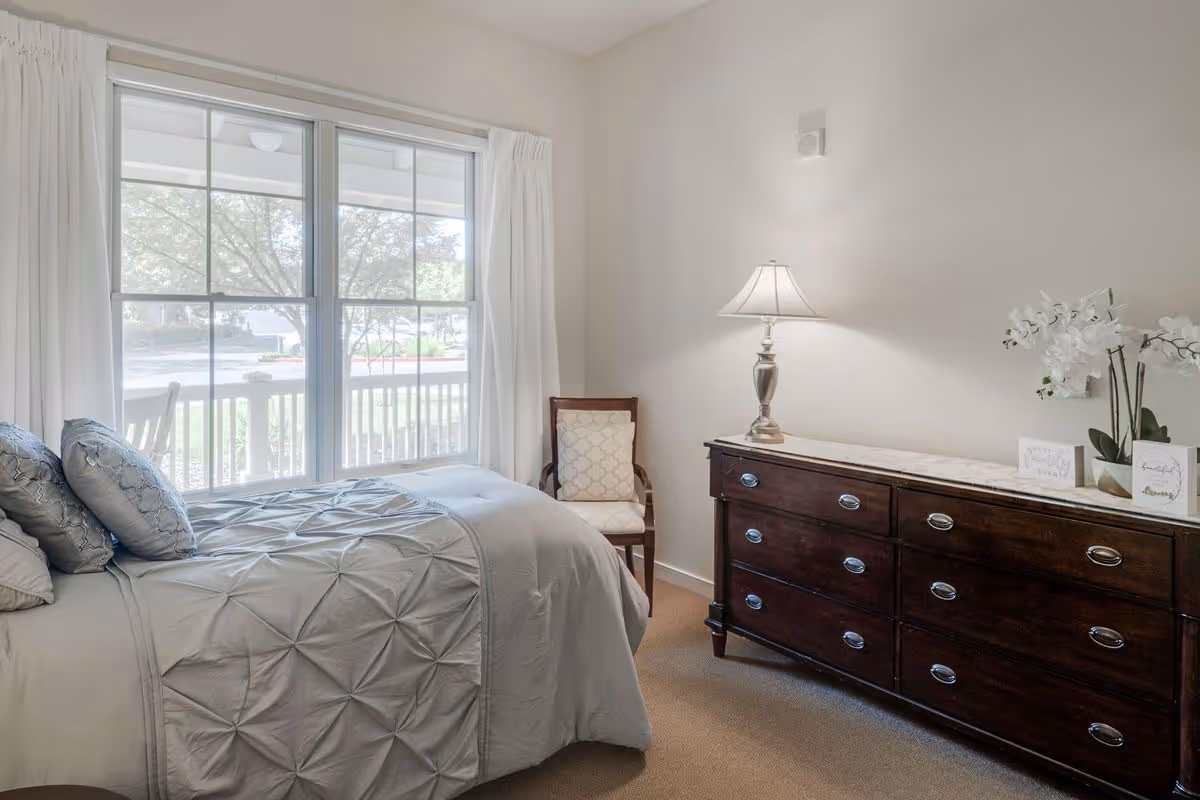 A bright bedroom with a large window covered by white curtains, a bed with gray bedding and pillows, a wooden chair with a patterned cushion, and a dark wooden dresser topped with a lamp and white orchid flowers.
