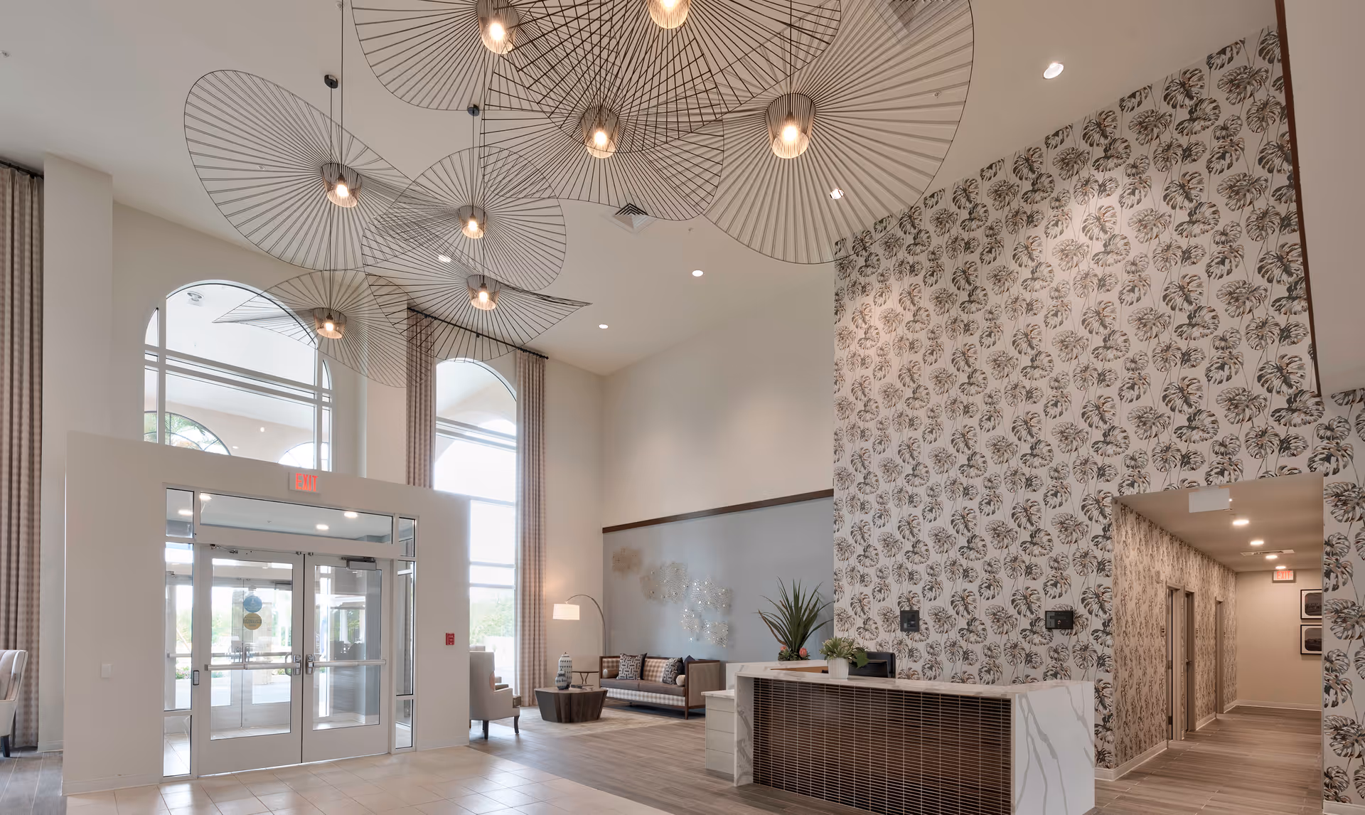 Spacious and modern assisted living facility lobby with high ceilings, large windows, decorative hanging light fixtures, a seating area with a sofa and armchair, a reception desk, and patterned wallpaper on one wall.