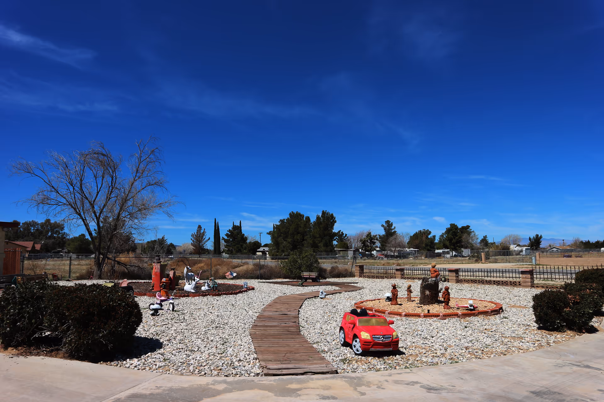 Outdoor garden area with a wooden pathway surrounded by gravel and small bushes. There are decorative statues and a small red toy car placed on the gravel. Trees and a clear blue sky are visible in the background.