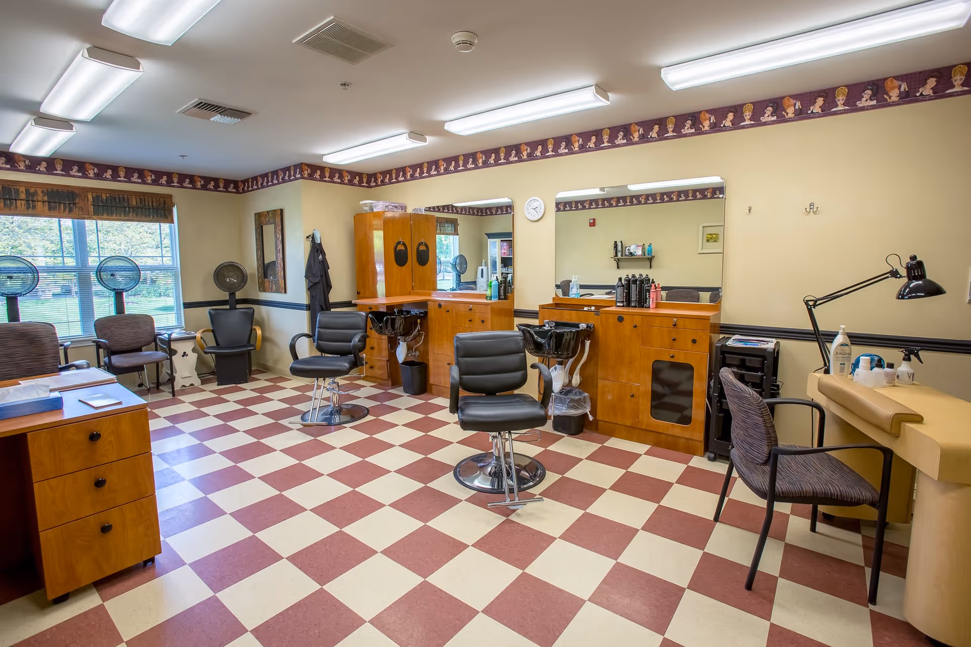 Interior view of a salon room with checkered red and white floor tiles, two black salon chairs in front of wooden counters with mirrors, hair care products on the counters, two black hair washing sinks, several chairs along the walls, and large windows letting in natural light.