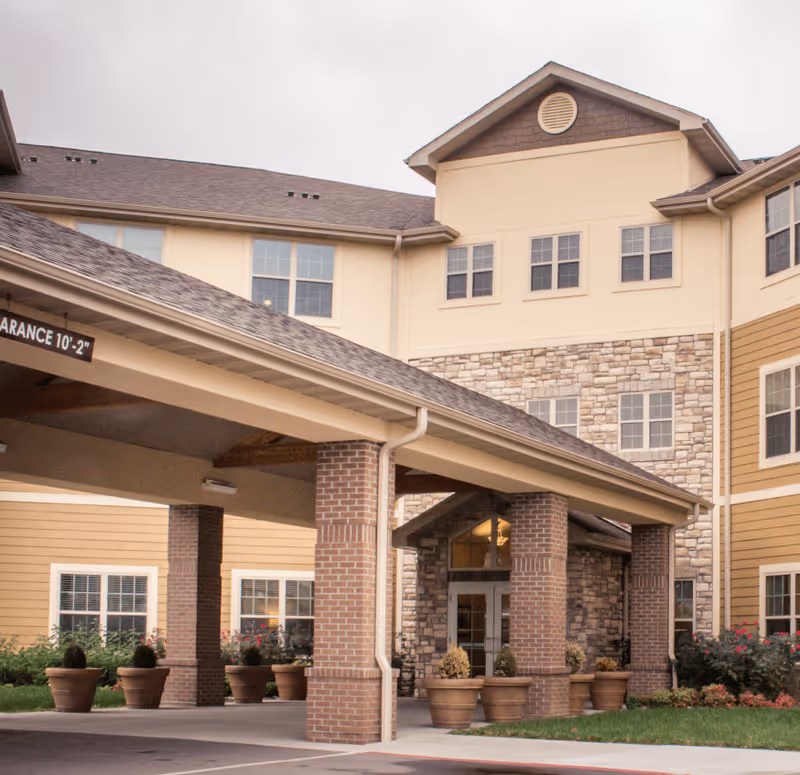 Exterior view of The Parkway Senior Living facility showing the entrance with a covered drop-off area supported by brick columns, large windows, and a combination of stone and siding on the building facade. Several large potted plants are placed near the entrance.