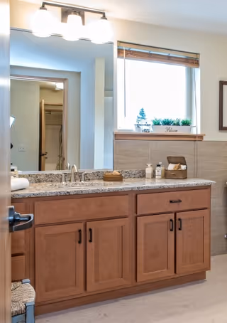 A bathroom with a granite countertop and wooden cabinets below. On the countertop are soap dispensers, a tissue box, and a small basket. Above the countertop is a large mirror with three light fixtures. A window with a wooden blind is above the sink, and a small plant and decorative sign are on the windowsill. The walls are tiled halfway up, and a door leading to another room is visible in the mirror reflection.