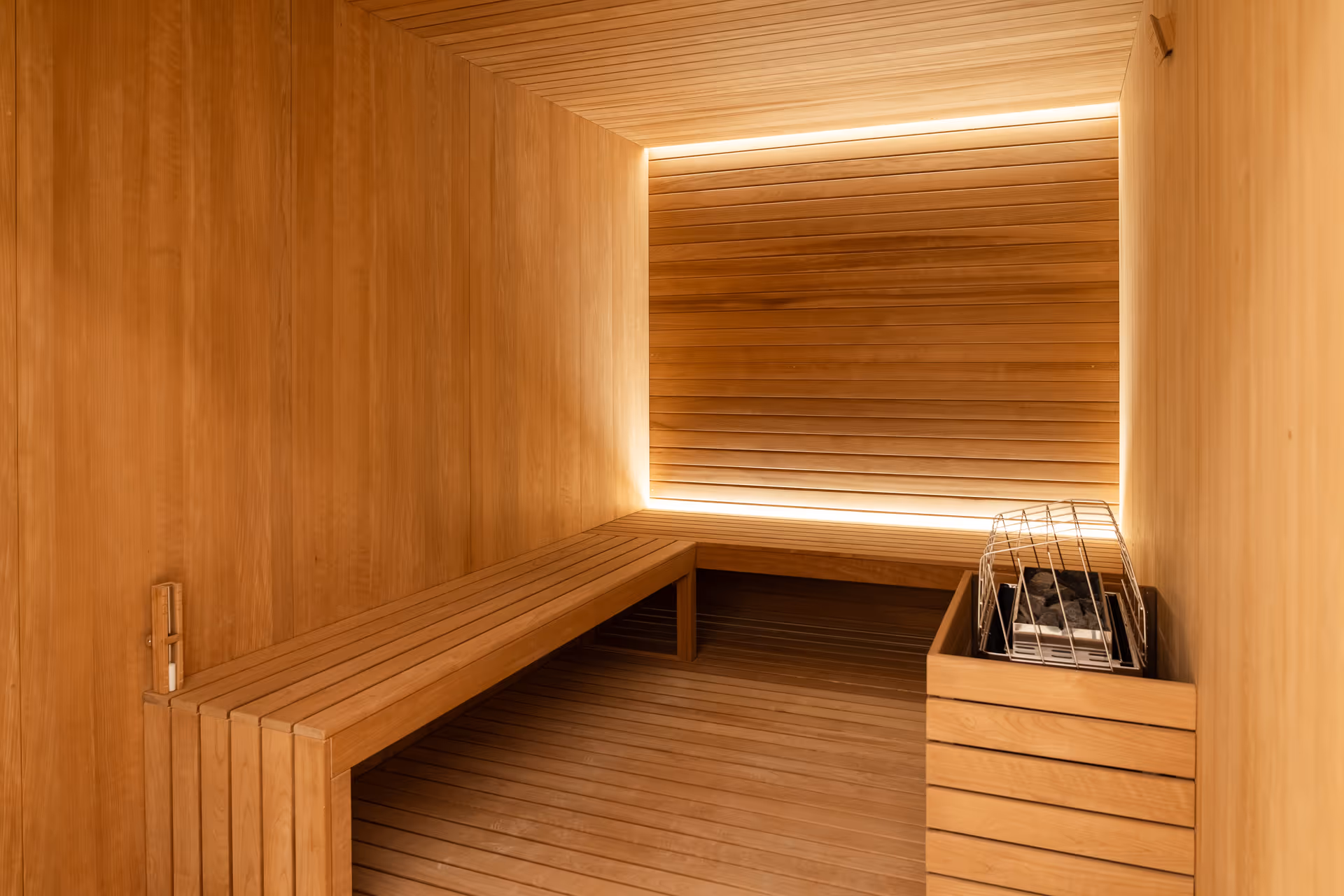 Interior of a wooden sauna room with a long wooden bench along one wall and a heater with stones in a metal cage on the right side. The walls, ceiling, and floor are all made of light-colored wood, and there is soft lighting behind a wooden panel on the far wall.