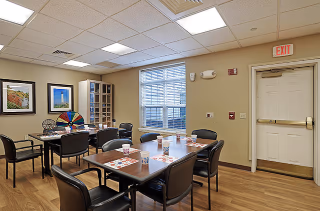 A well-lit room with two rectangular tables arranged for a game or activity, each table surrounded by black chairs. On the tables are bingo cards, cups, and a colorful spinning wheel. The room has wooden flooring, beige walls, a window with blinds, framed pictures on the wall, and a white exit door.