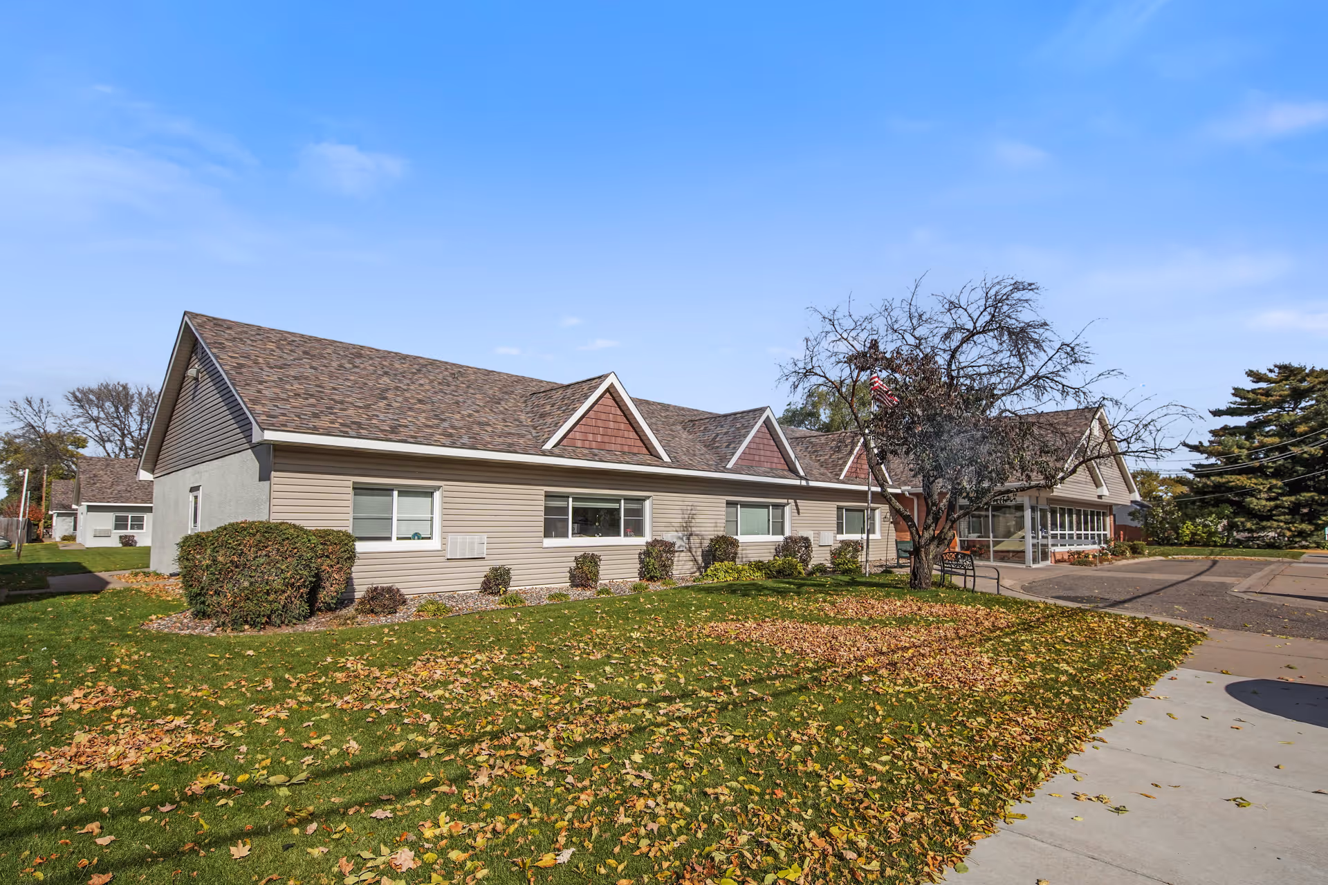 Exterior view of Osseo Gardens Assisted Living facility showing a single-story building with beige siding and a brown shingled roof. There is a tree with sparse leaves and an American flag near the entrance. The lawn has scattered fallen leaves and a sidewalk runs alongside the building.