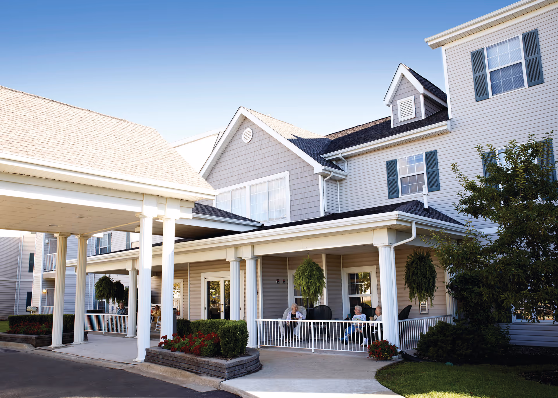 Exterior view of a senior living facility with a covered entrance and porch area. Three elderly people are sitting and conversing on the porch, which is decorated with hanging plants and flower beds. The building has light-colored siding, multiple windows with shutters, and a clear blue sky overhead.