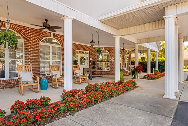 Covered outdoor patio area with white columns, wooden rocking chairs with cushions, hanging plants, and red flowers along the edge of the concrete walkway in front of a brick building with large windows and a ceiling fan.