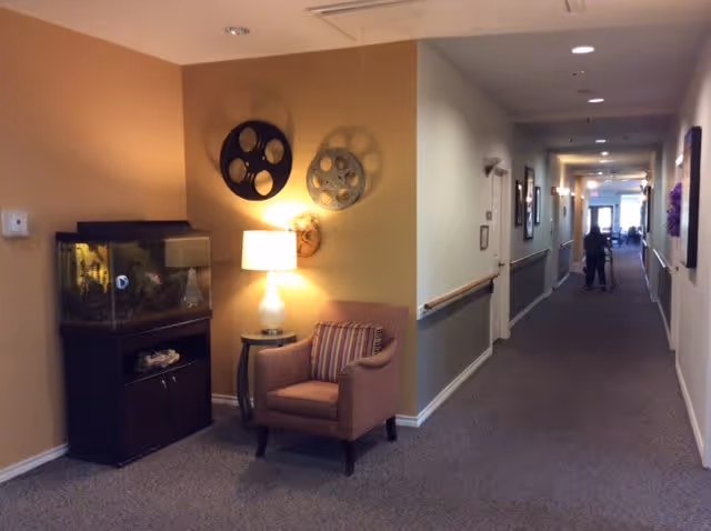 A hallway in a senior living facility with a carpeted floor and handrails along the walls. On the left side, there is a small seating area with a striped armchair, a side table with a lamp, and a fish tank on a dark wooden stand. The wall behind the seating area is decorated with three large film reel wall hangings. At the far end of the hallway, a person is visible walking with a walker.
