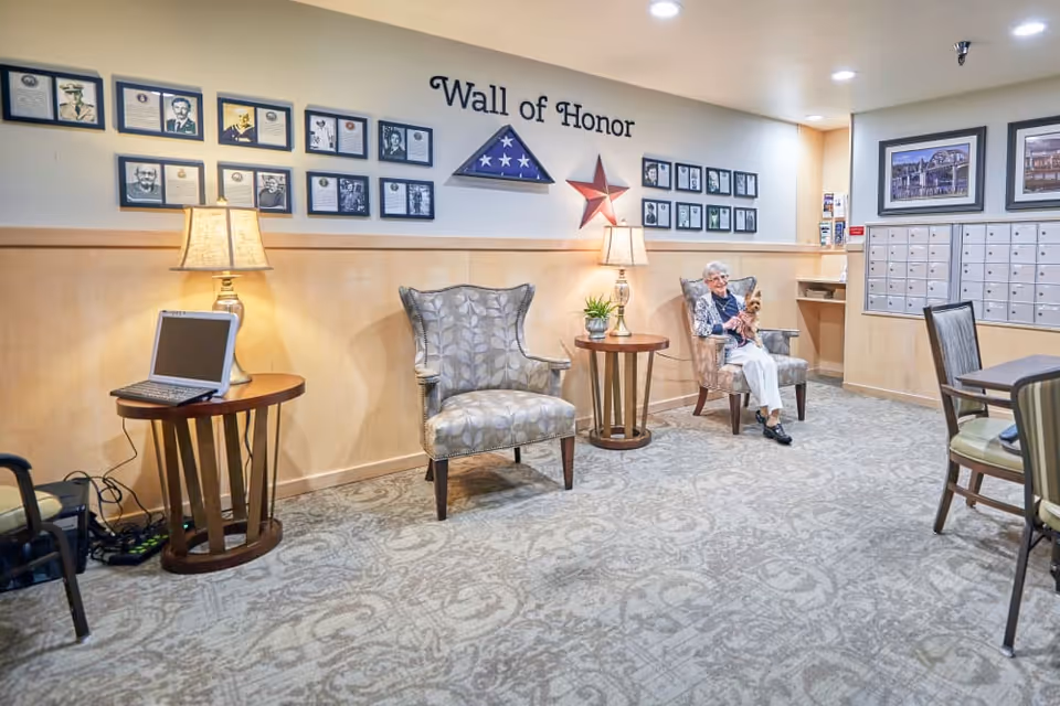 A cozy seating area in a senior living facility with two patterned armchairs, two side tables with lamps, and a laptop on one table. An elderly woman is sitting in one chair holding a small dog. The wall behind them features a 'Wall of Honor' display with framed photos and a folded American flag in a triangular case. There are also mailboxes and framed pictures on the adjacent wall.
