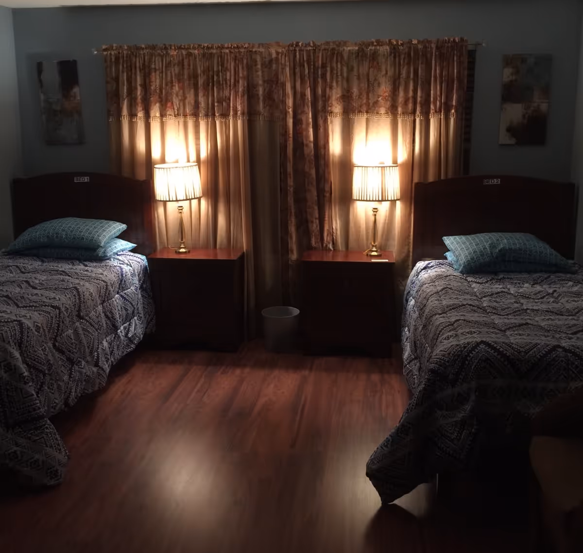 A dimly lit bedroom with two single beds labeled BED 1 and BED 2. Each bed has patterned bedding and two pillows. Between the beds are two wooden nightstands, each with a lit table lamp. Behind the nightstands is a window covered with floral curtains. The floor is wooden, and there is a small trash bin between the nightstands.