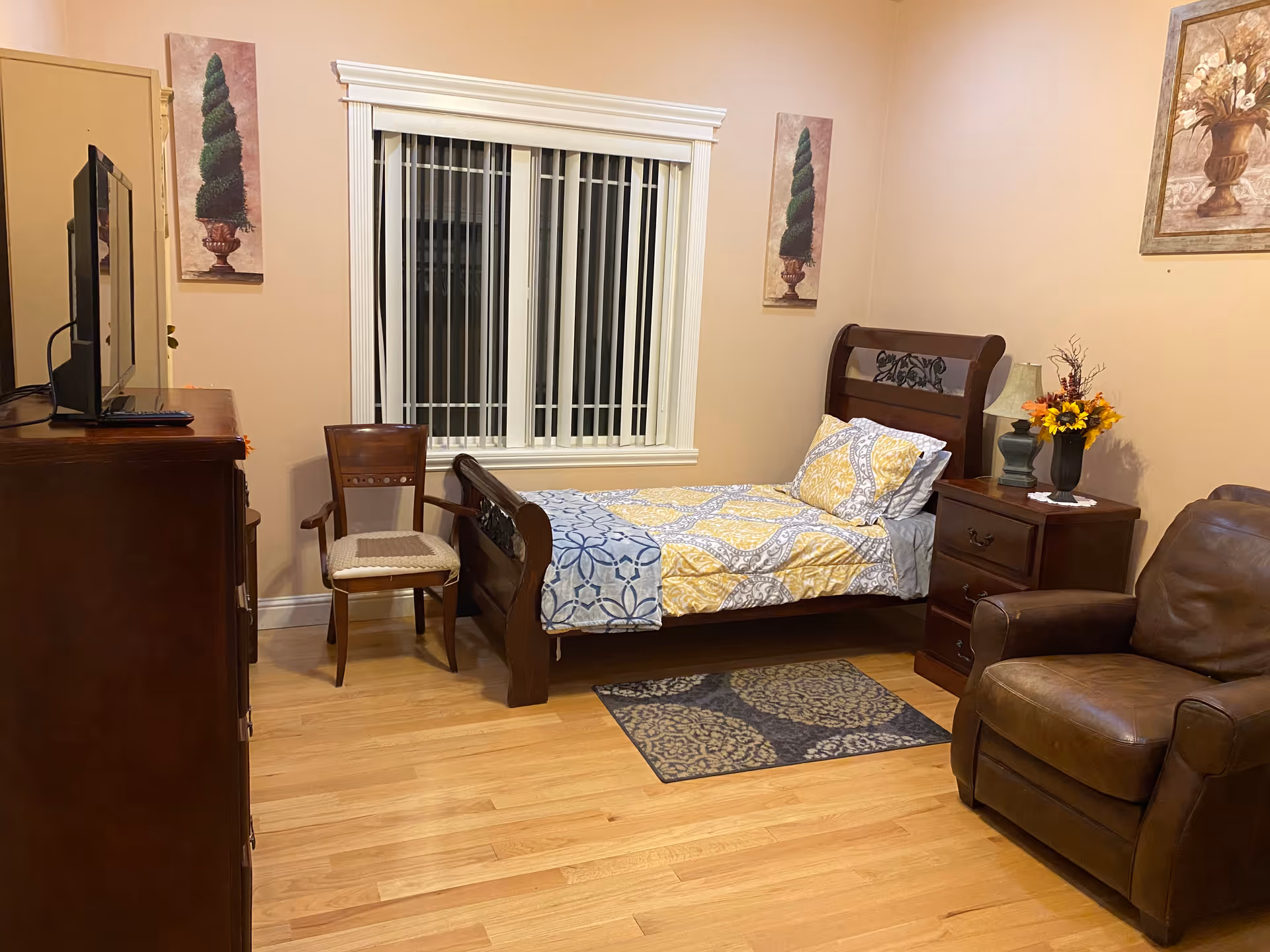 A cozy bedroom with a single wooden bed featuring yellow and gray patterned bedding. Next to the bed is a wooden nightstand with a lamp and a vase of flowers. A brown leather armchair is positioned near the nightstand. Across from the bed is a wooden dresser with a flat-screen TV on top. A wooden chair with a cushion is placed between the dresser and the window. The walls are painted beige and decorated with framed artwork. The floor is light wood with a small patterned rug near the bed.
