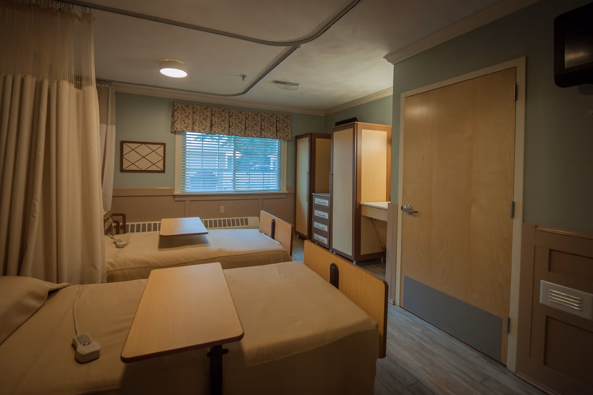 A room with two hospital-style beds separated by a curtain. Each bed has a wooden overbed table and a remote control. The room has a window with blinds and a floral valance, a wooden door, and a set of wooden cabinets and drawers. The walls are painted in muted tones and the floor has a wood-like finish.