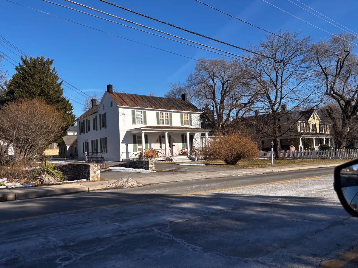 Two-story white house with a front porch and neighboring homes along a street under a clear blue sky.