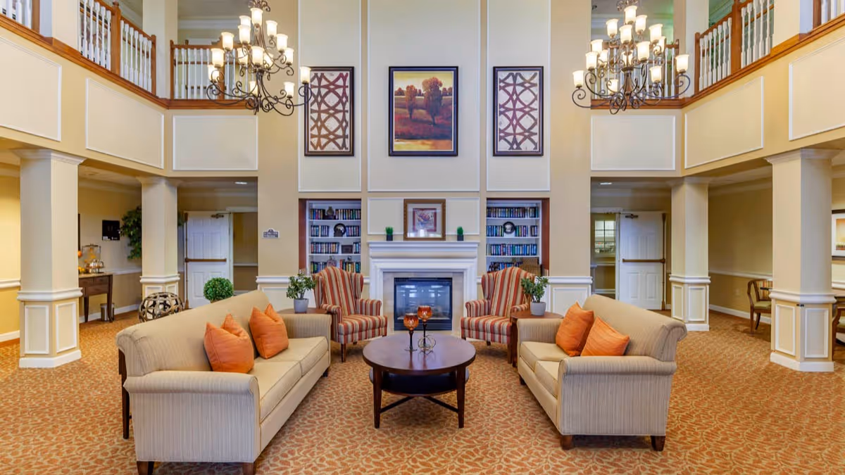 A spacious and elegant living room area in a senior living facility with two beige sofas adorned with orange pillows, two striped armchairs, a round wooden coffee table, and a fireplace with built-in bookshelves on either side. The room features high ceilings with two chandeliers, framed artwork above the fireplace, and a carpeted floor with a patterned design.