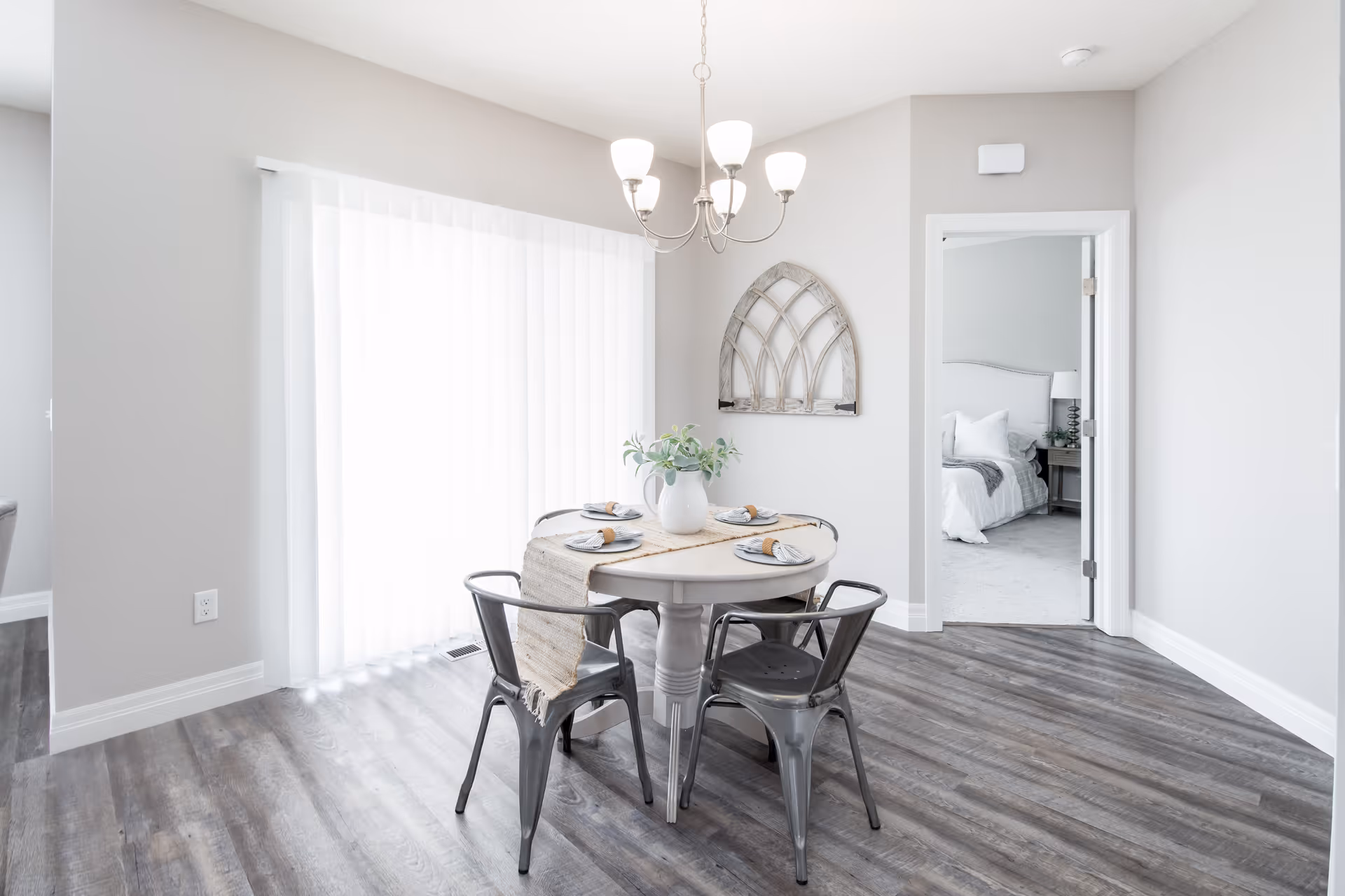 A bright dining area with a round table set for four, featuring a beige table runner and a white vase with greenery. Four metal chairs surround the table. A chandelier with five lights hangs above. Sliding glass doors with vertical blinds allow natural light to fill the room. A doorway leads to a bedroom with a neatly made bed and a bedside table with a lamp. The flooring is a dark wood laminate, and the walls are painted light gray.