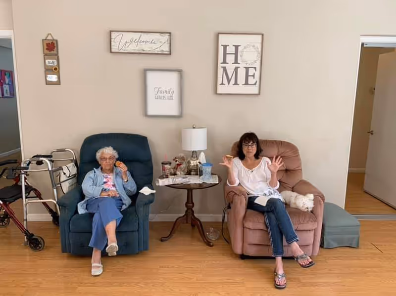 Two women sitting in comfortable armchairs in a living room area. The woman on the left is elderly with white hair, wearing glasses, a light blue jacket, and blue pants, holding a snack. The woman on the right has dark hair, glasses, a white blouse, and jeans, gesturing with her hands. Between them is a small round table with a lamp, cups, and other items. Wall decorations include framed signs with the words 'Welcome', 'Family Gathers Here', and 'HOME'. A walker is visible to the left of the elderly woman, and a small white dog is lying on the armchair next to the woman on the right. The floor is wooden, and there is an open doorway to the right.