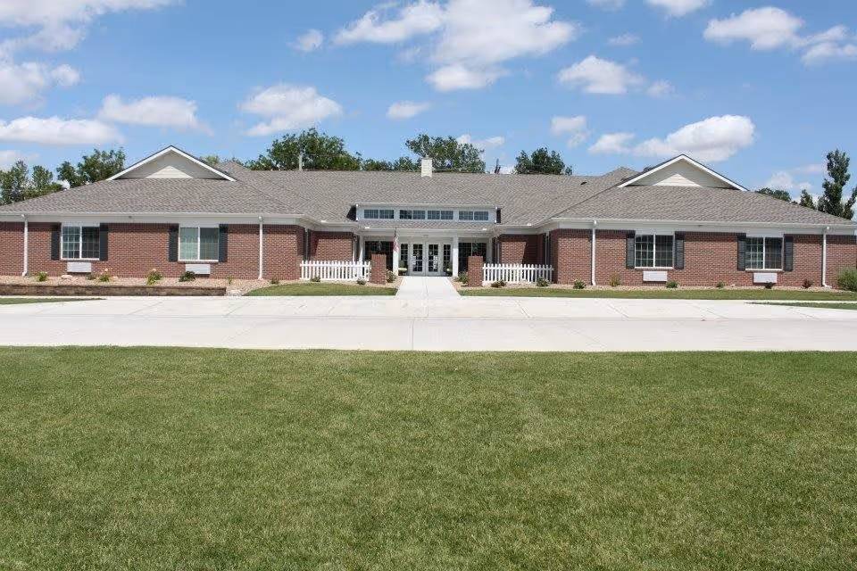 Front exterior view of a single-story brick building with a gray shingled roof, white trim, and multiple windows. There is a concrete walkway leading to the main entrance, which has glass double doors and a small white picket fence on either side. The building is surrounded by a well-maintained green lawn and some small shrubs, under a partly cloudy blue sky.