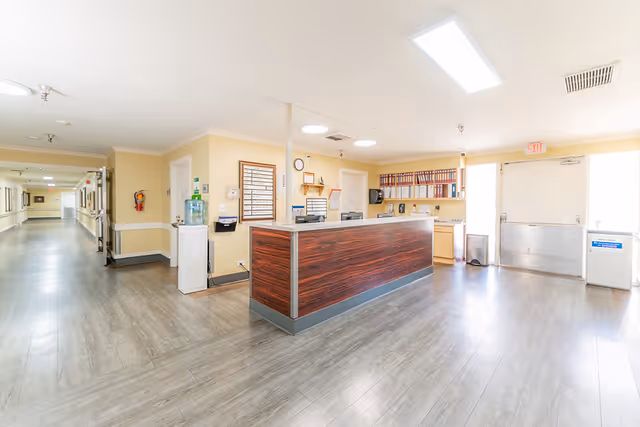 Bright and clean reception area in a healthcare facility with a wooden front desk, water cooler, filing shelves with binders, and a long hallway extending to the left.