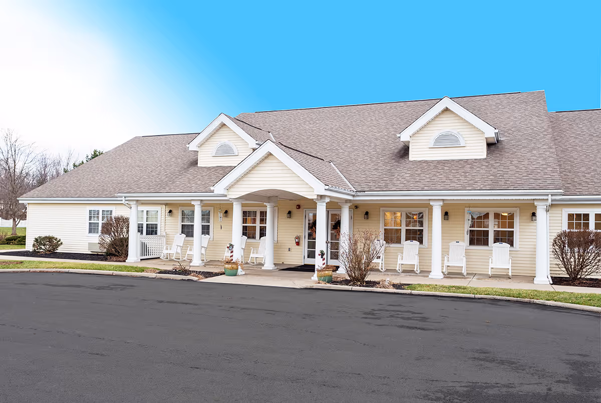 Front exterior view of Barnes Place facility showing a single-story building with beige siding, white columns, several windows, and white rocking chairs on the porch. The sky is clear and blue.