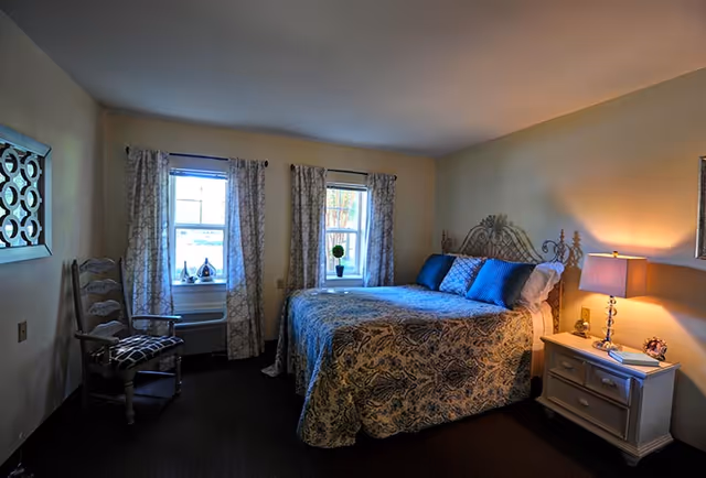A cozy bedroom with a large bed covered in a patterned blue and beige bedspread, three blue pillows, and a decorative metal headboard. Two windows with patterned curtains allow natural light into the room. A wooden chair with a patterned cushion is placed near one window. A white nightstand with two drawers holds a lamp, a small clock, and a decorative item. The walls are painted a soft beige color.