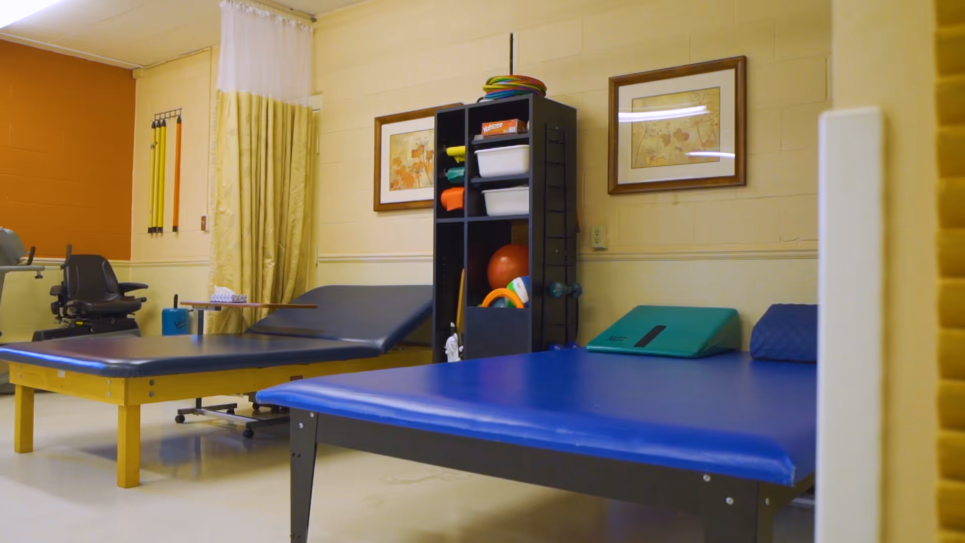 An interior therapy room with blue treatment tables, exercise mats, a shelving unit holding therapy equipment, and framed artwork on the wall.