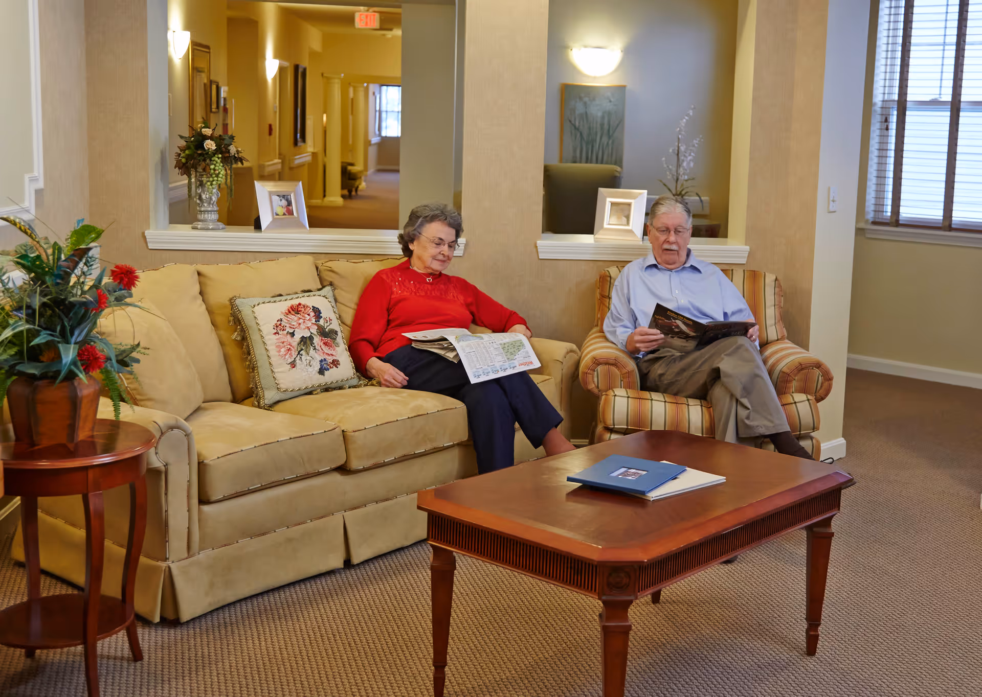 An elderly woman and man sitting in a cozy living room area. The woman is seated on a beige sofa with floral pillows, reading a newspaper, while the man is seated on a striped armchair reading a magazine. A wooden coffee table with books is in front of them, and there are decorative plants and framed pictures on the walls and surfaces. The room has warm lighting and a window with blinds.