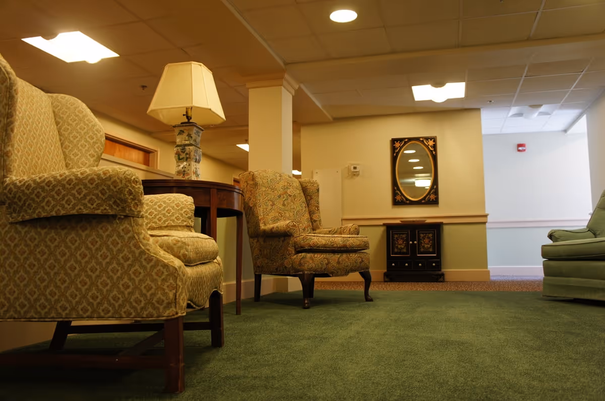 Low-angle view of a furnished seating area with upholstered armchairs, a side table with a lamp, and a wall mirror in a carpeted common room.
